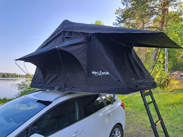 Black Silma rooftop tent mounted on white car by lakeside with forest background